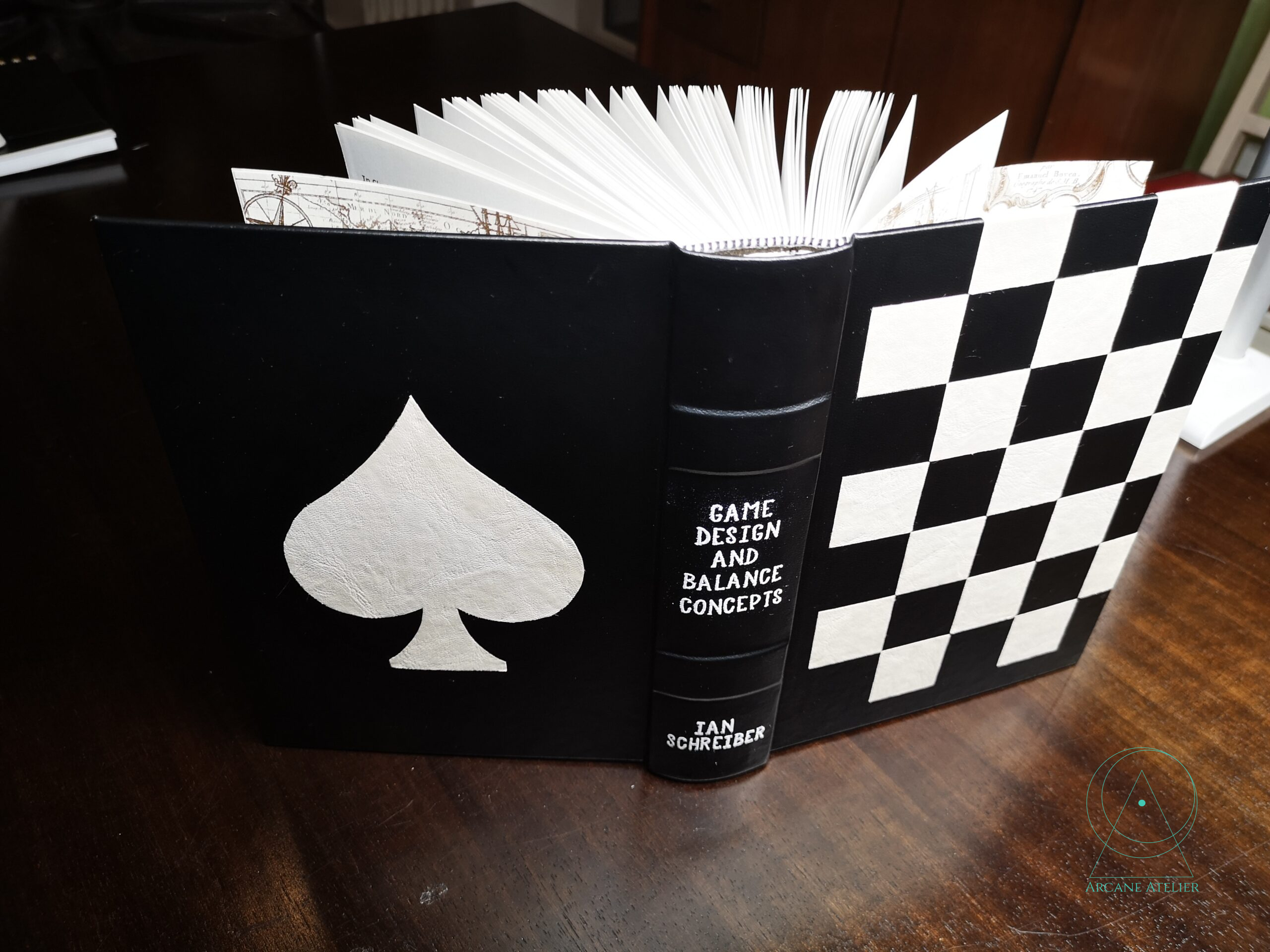 Standing, fully open view of the Game Concepts Book showing a brown faux-leather cover with stitched chessboard pattern and a white spade symbol on black on the back cover.