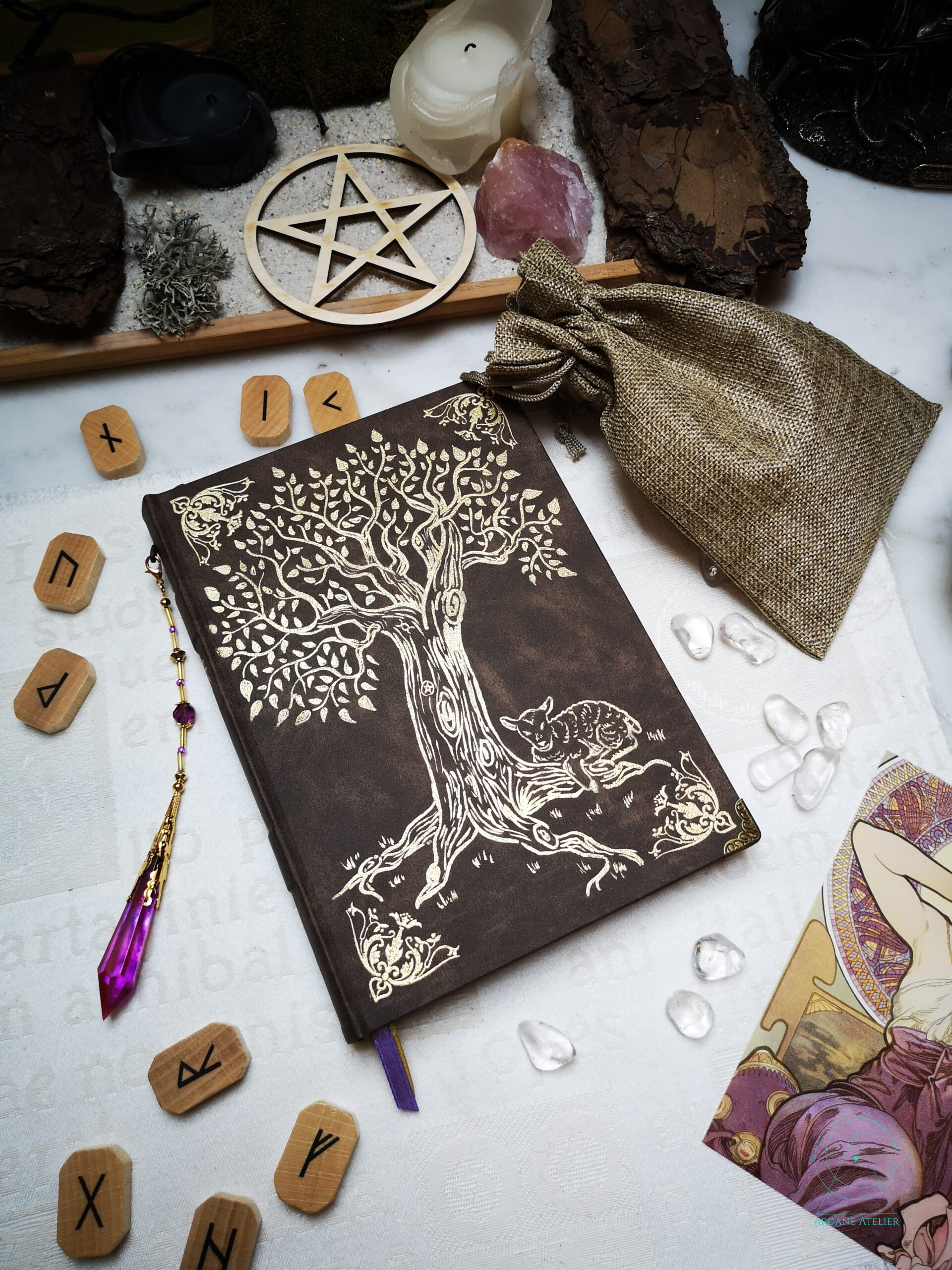 Brown leather journal with a gold-embossed tree and a small lamb at its base, featuring a violet crystal pendant on a gold chain.