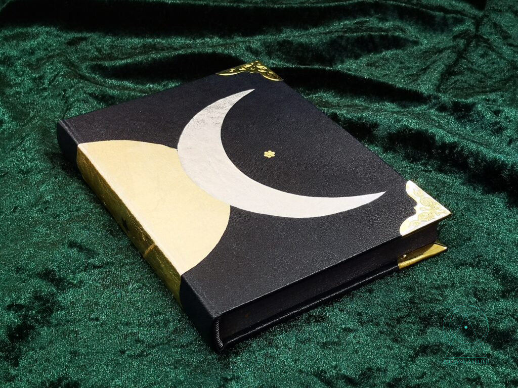 Three-quarter view of a black leather journal showing part of the triple moon motif with the crescent and half of the full moon.