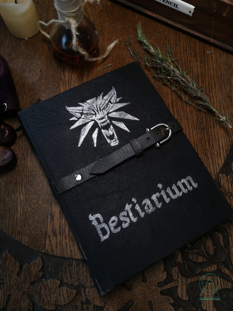 Black aged-leather journal with a belt closure, featuring the silver Witcher wolf emblem and the word ‘Bestiarium’ on the cover.