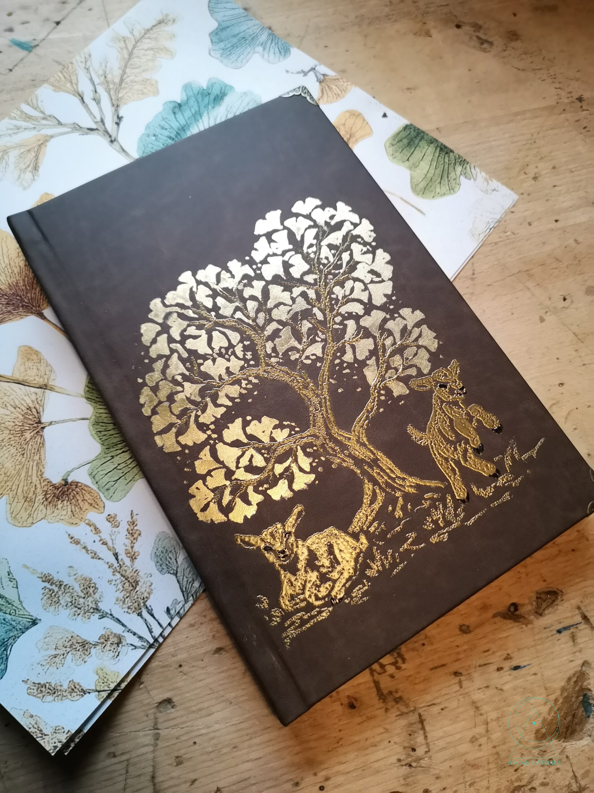 Brown journal with a gold-embossed ginkgo tree and gilded leaves, featuring two small goats at the base of the trunk.