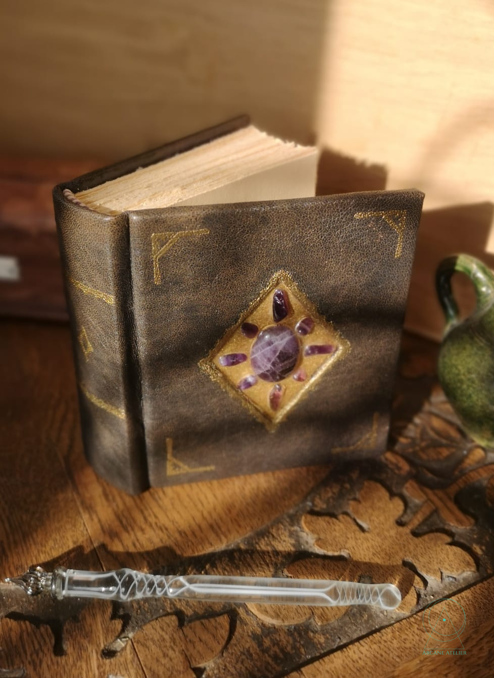 Three-quarter view of a square brown leather journal showing the thick cream paper block and the central amethyst inlay set in a gold diamond frame.